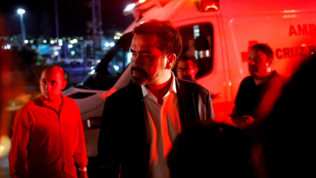 Man in a Dark Suit Stands Near a Red-lit Ambulance at Night, with People and Emergency Activity in the Background - Regios En México