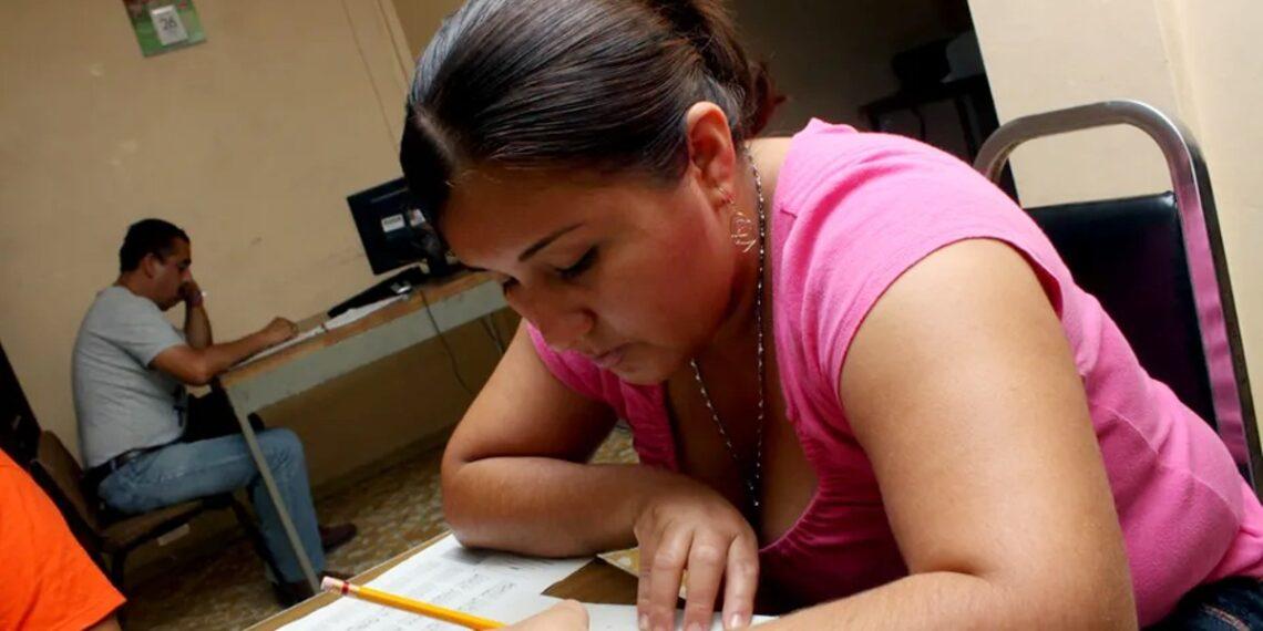 Woman in a Pink Top Leans over a Desk, Writing on Papers As a Man Sits at a Computer in the Background. - Regios En México