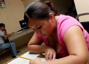 Woman in a Pink Top Leans over a Desk, Writing on Papers As a Man Sits at a Computer in the Background. - Regios En México