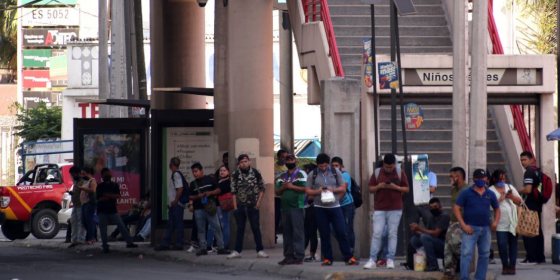 People Waiting in a Queue Along a City Sidewalk Beside a Bus Shelter and Concrete Stairs, with a Red Emergency Vehicle Nearby. - Regios En México