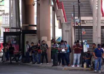 People Waiting in a Queue Along a City Sidewalk Beside a Bus Shelter and Concrete Stairs, with a Red Emergency Vehicle Nearby. - Regios En México