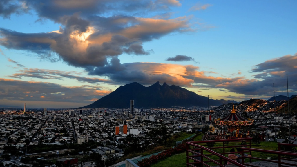 Por-que-congelaron-el-dinero-de-los-municipios - Regios En México Sunset over a City with a Prominent Mountain in the Distance and a Lit Carousel in the Foreground, with a Railing and Flowers Nearby. - Regios En México